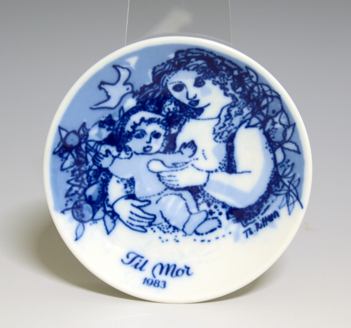 Morsdagsplatte av porselen. Hull til oppheng bak. Hvit glasur. Dekorert i blått med sittende kvinne som mater et lite barn, tekst Mors Dag 1986.

