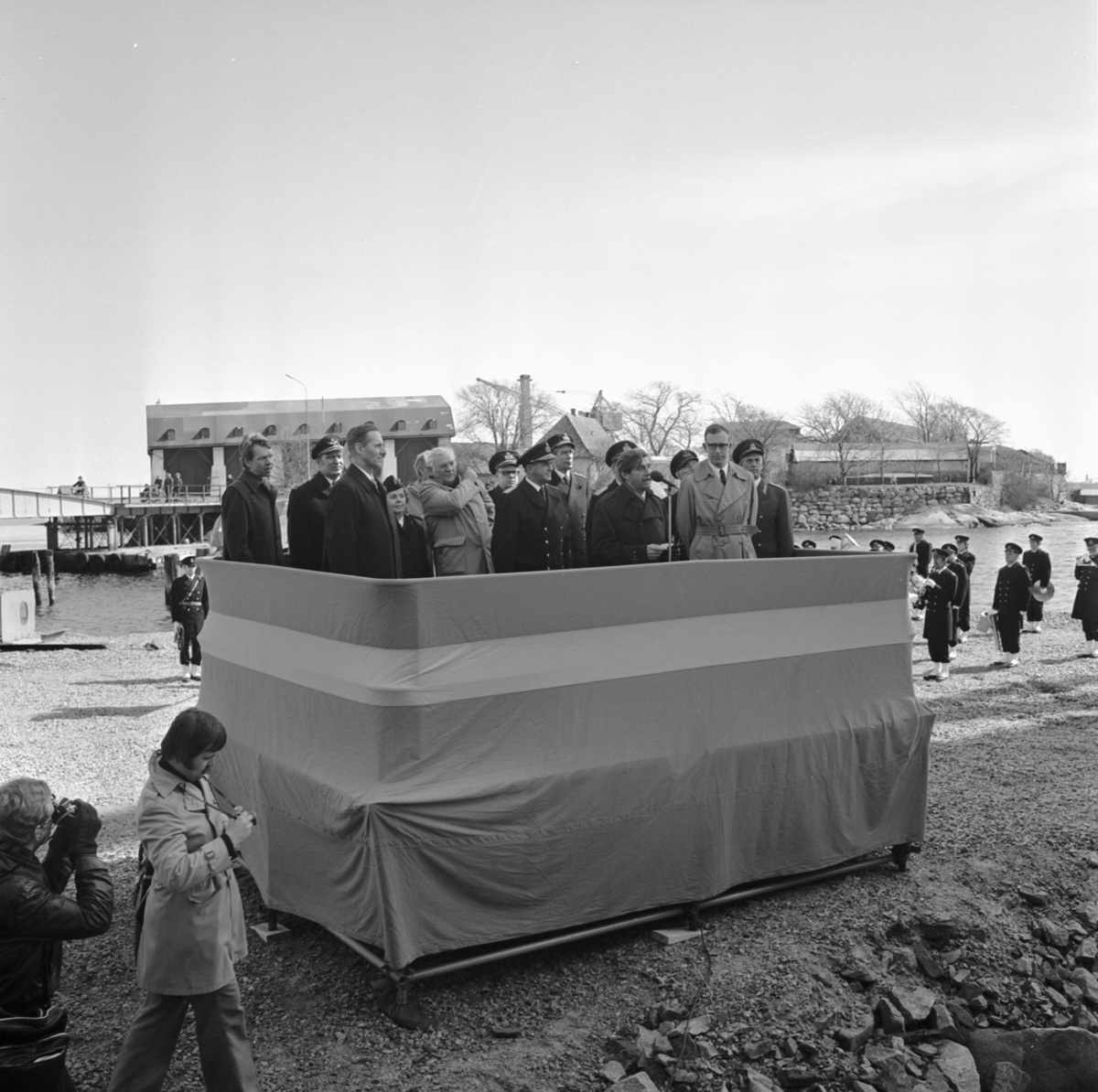 På en podium står en munter skara höga militärer och politiker som håller tal under sjösättningen av T 141. I bakgrunden syns Wasaskjulet och Lindholmsbron.