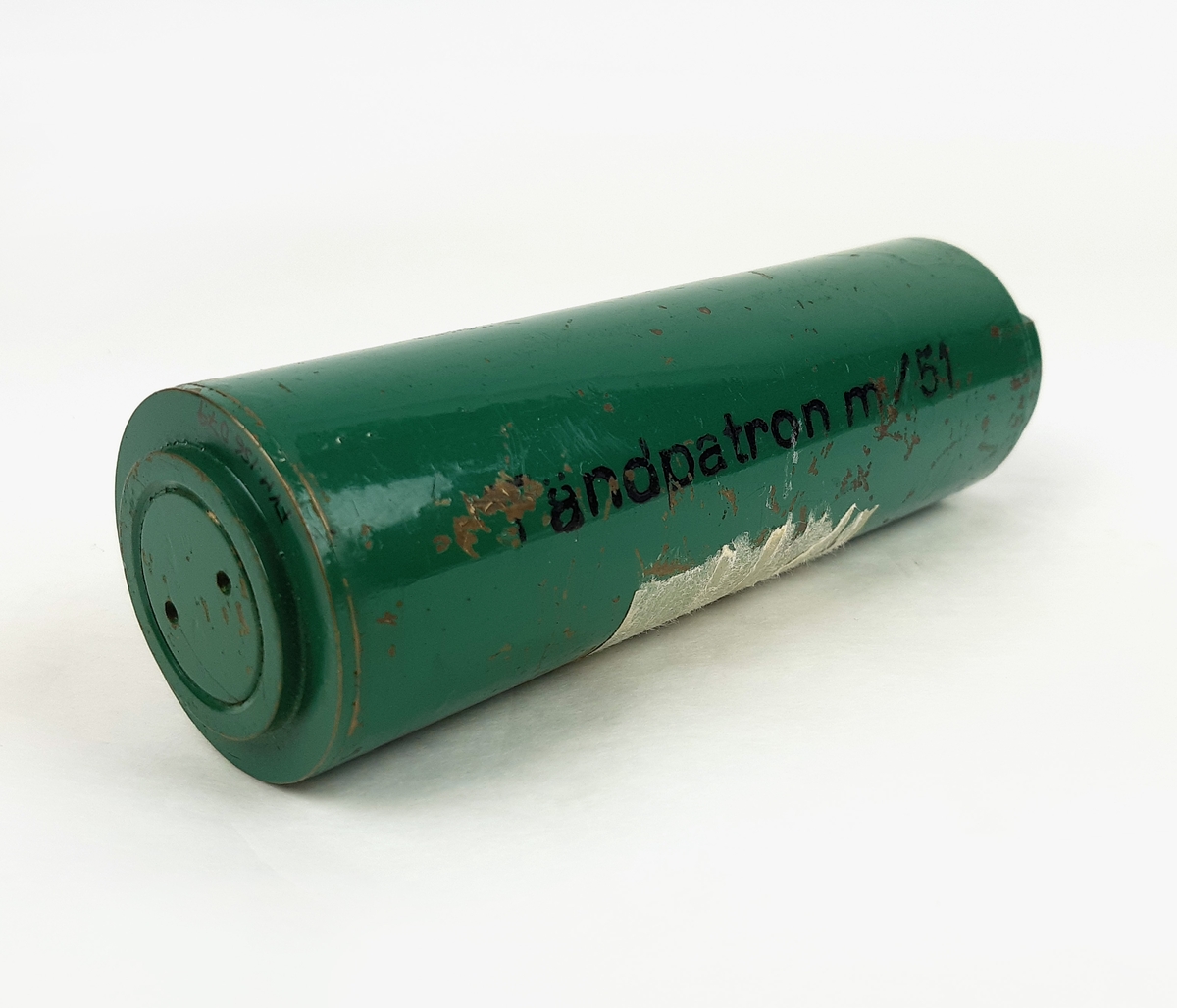 Tändpatron m/51, bestående av en cylinderformad kapsell i grönmålad metall. Vid sidan märkt "Tändpatron m/51". Samt märkt "BLIND" på kortsidorna.