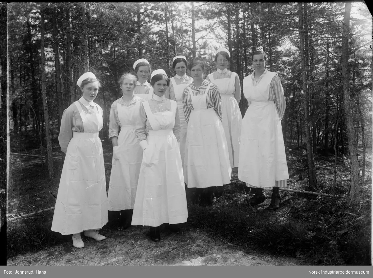 Gruppeportrett av åtte stående kvinner i sykepleier uniform foran skog. Søster Constanse står fremmerst i midten.