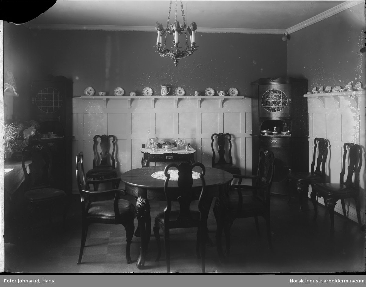 Interiør av spisestue. Tre stoler står rundt et spisebord midt i rommet. Fem stoler til er synlige langs veggene. Pyntetallerkener og -kar står på hyller langs veggene. I de to synlige hjørnene av rommet står det hjørnekabinett. Et bord med sølvtøy er synlig langs bakre vegg.