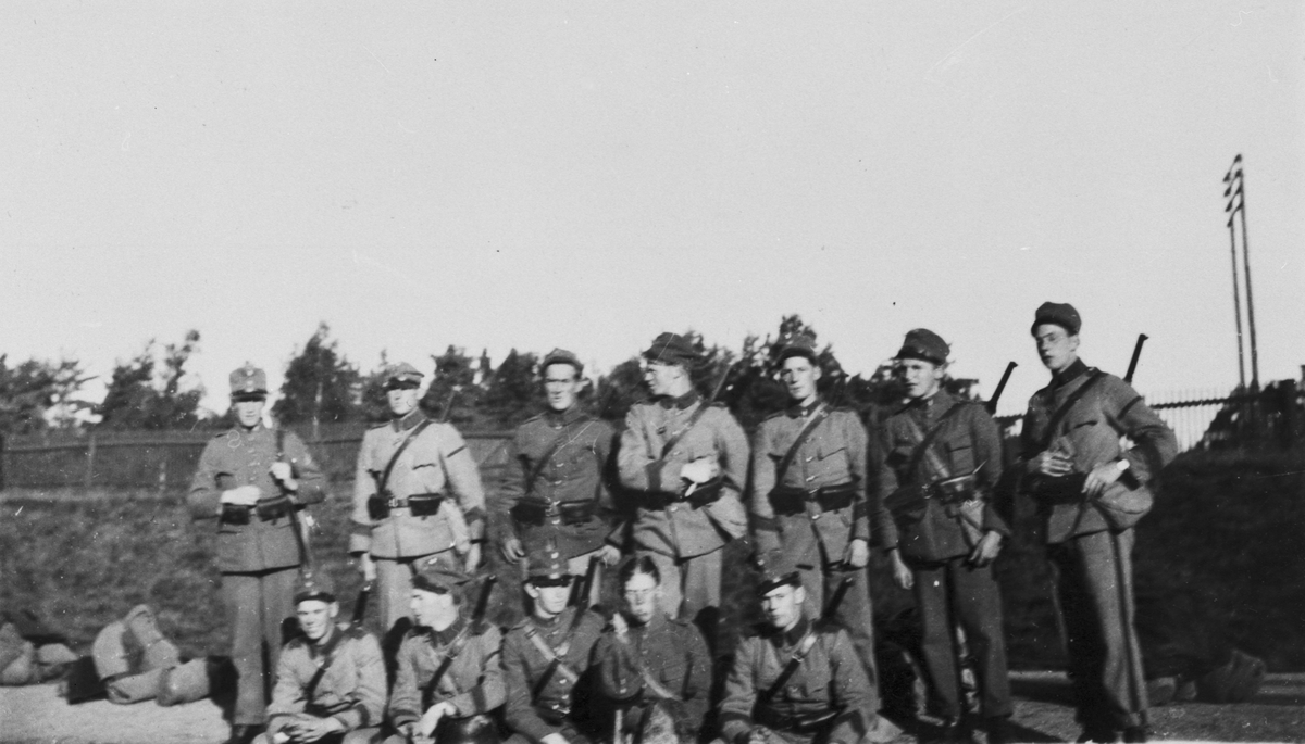 Gruppfoto av tolv militärer utrustade med gevär vid ett staket och med packade väskor, ca 1927.