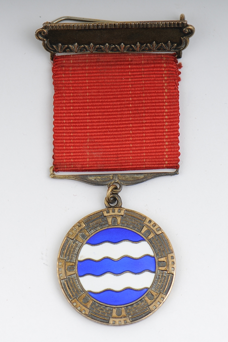 Medalje med stillisert Harstad byvåpen, bymur med tårn og bølger, festes med nål, bladkrans på nåldel.