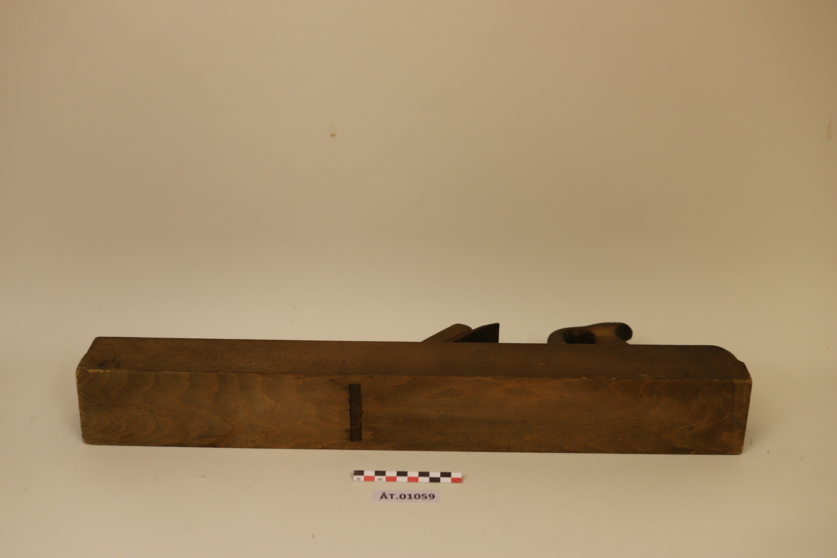 Rektangulær langhøvel med jern, kile og bøylehåndtak i bakenden. Høvelen er avrundet bak og skåret inn i hjørnene fremme