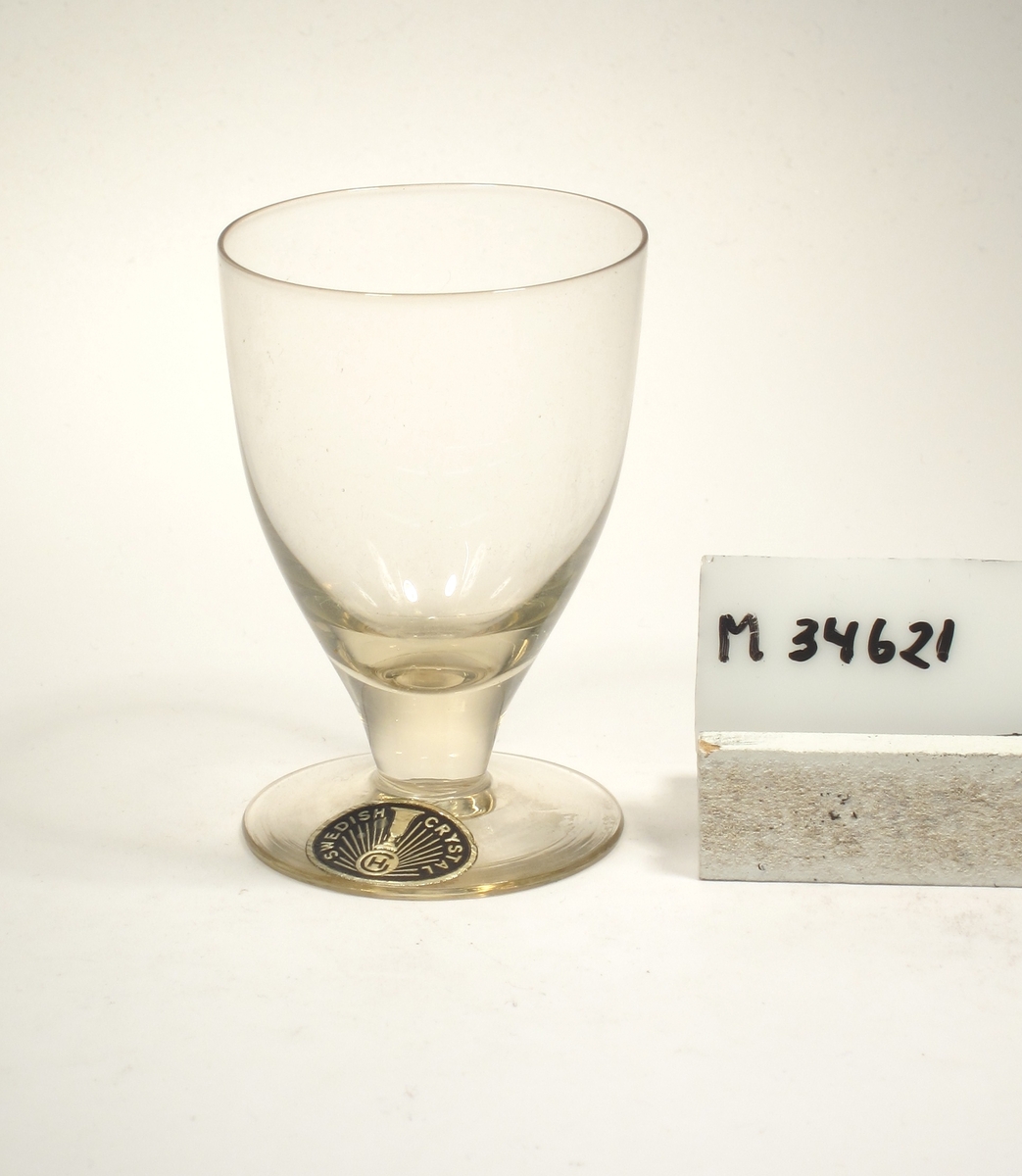 Gultonat. Ovoid kupa med draget tjockt konande ben.
Etikett: Oval, svart botten med guldtext: SWEDISH CRYSTAL, GH Strålar och ett glas.