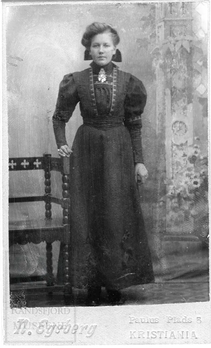 Kvinne i helfigur stående i mørk kjole med høy hals og puffermer.