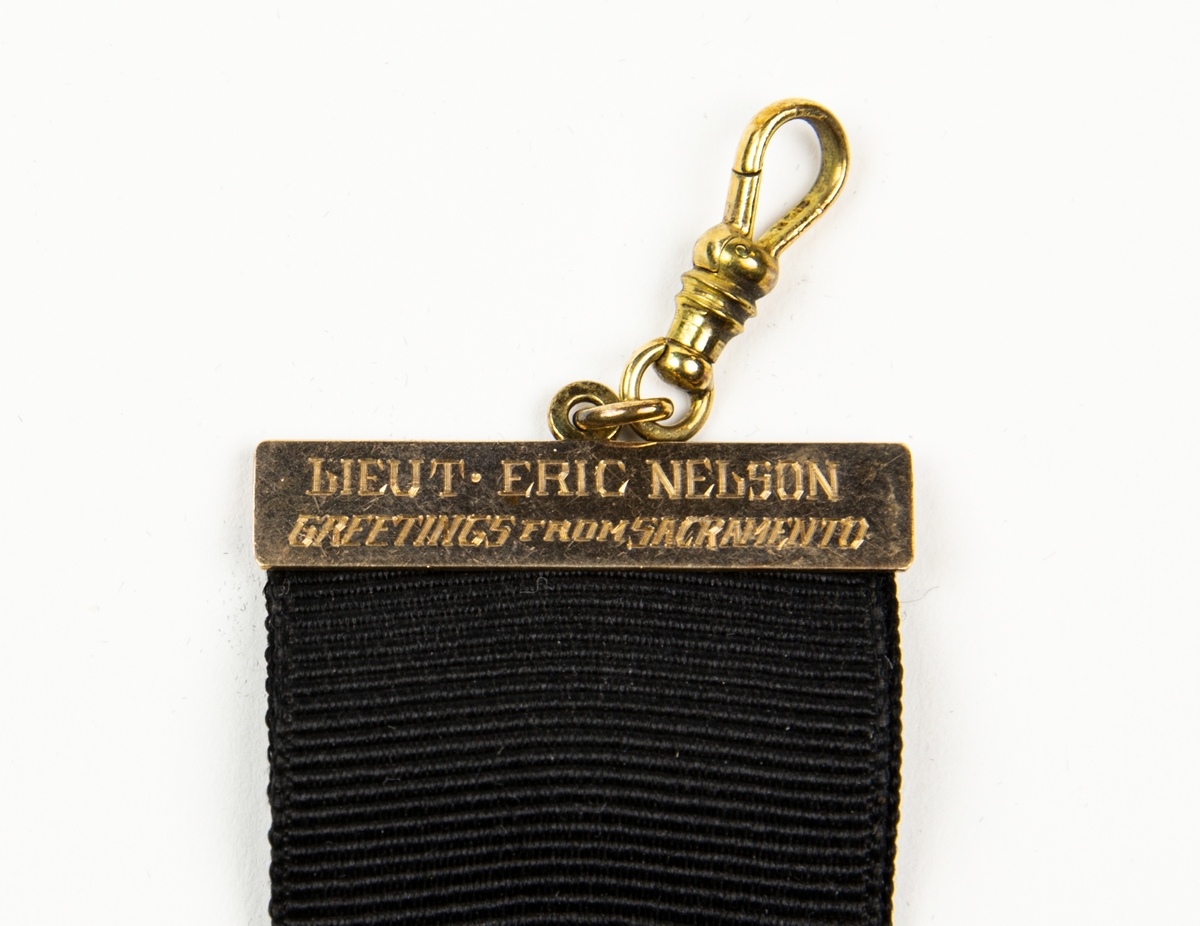 Medaljong med guldstroff. Medaljongen består av glas med en ram av guld. Medaljongen hänger i ett svart medaljband som i överkant har ett fäste med texten "LIEUT ERIC NELSON GREETINGS FROM SACRAMENTO".