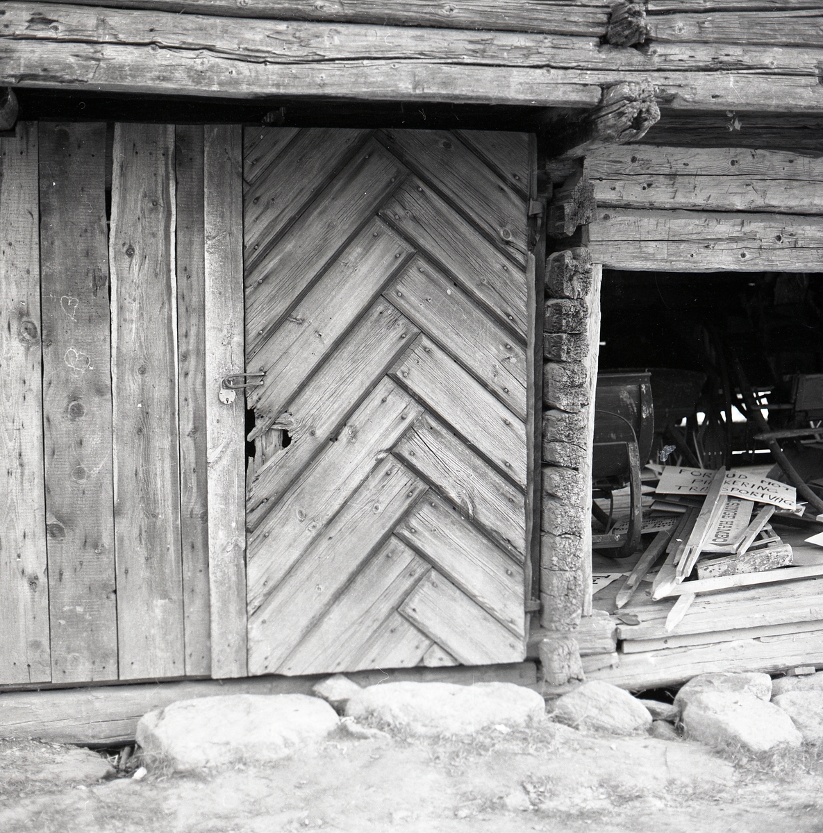 Trädörr med hänglås på en gammal timmerbyggnad i Arbrå. Bredvid dörren syns ett förråd med bland annat några skyltar.
