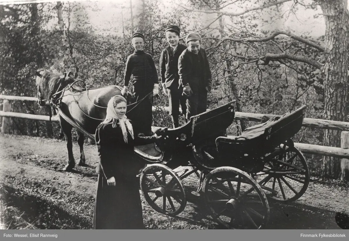 Tre smågutter står på en hestevogn og en kvinne står ved siden av vognen. Ellisif Wessel har tatt dette bildet langs veien på strekningen mellom Strand (ved Asmund Ryeng?) og Svanvik (ved Salmovaara?). 