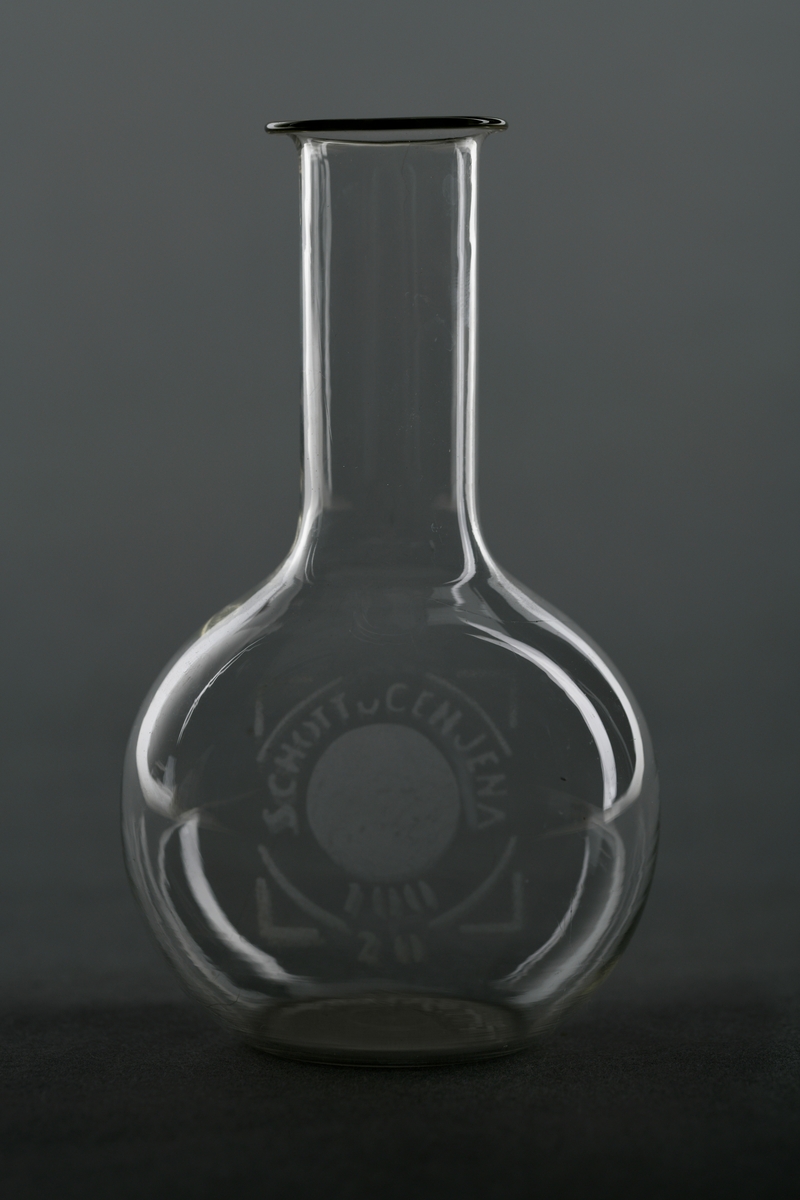 En målekolbe av glass, trolig borosilikatglass. Glasskolben har rund form med lang og relativt tynn hals. Ofte blir slike kolber brukt til å lage løsninger med et visst volum og konsentrasjoner.
