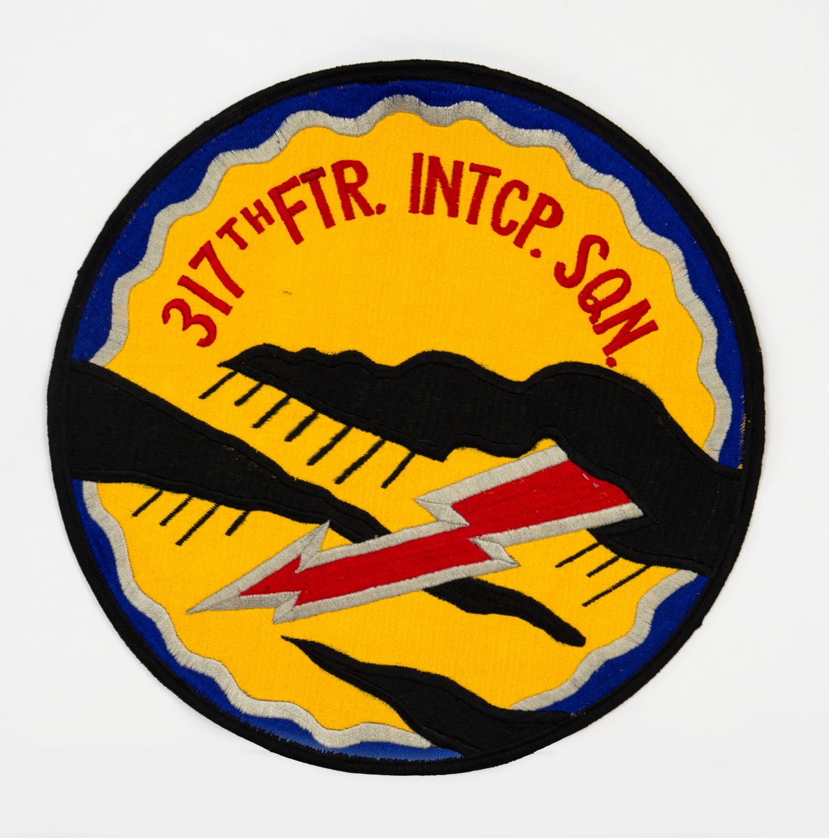 Broderat emblem på gul botten. Motiv med två svarta regnmoln och en röd blixt. Text: "317TH FTR. Intcp. Sqn" (317th Fighter Interceptor Squadron).