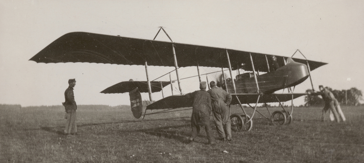 Flygplan SW 10 Farman med motor igång står på flygfältet på Malmen, cirka 1914-1915. Flygförare i flygplanet och personal ur Flygkompaniet i arbete bredvid.