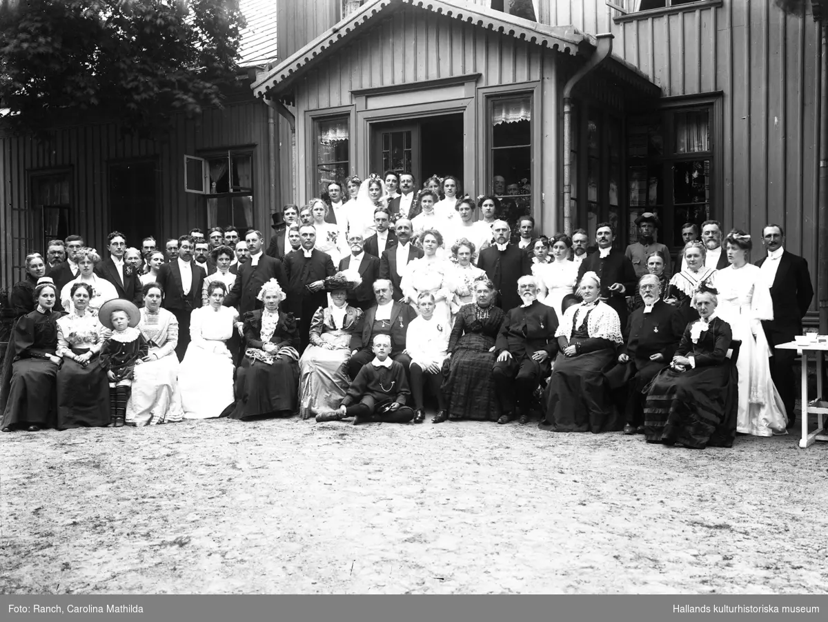Karin Holmdahl, bröllop Tvååker. Gruppbild av brudpar med ett stort antal bröllopsgäster, fotograferade framför ett bostadshus. I bakre raden till höger står en yngling i uniform.