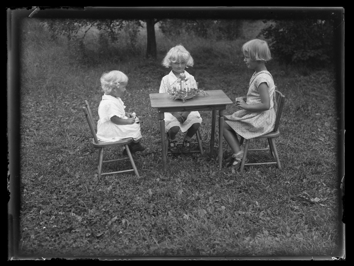Tre småflickor sitter på barnstolar vid bord med leksak på i trädgård. Västerås.