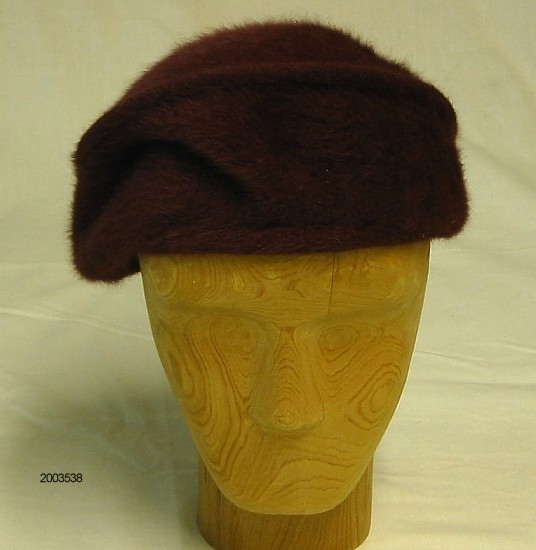 Hatt. Fiolett hatt med rund pull og uten brem. Søm rundt hele formen markerer toppen. Stoffet er mykt og flosset. Innvendig er hatten kantet med et strikk. Videre er det sydd i et svart hattestrikk og  produksjonmerke.