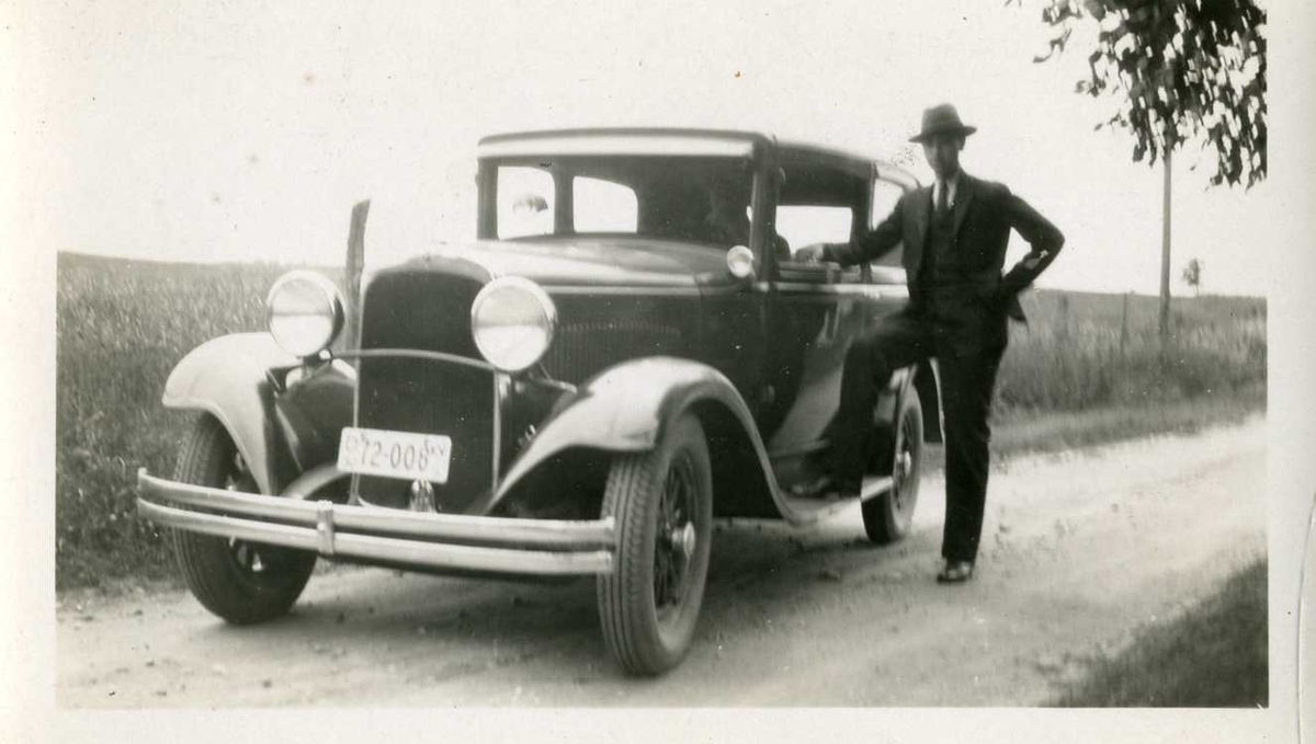 Olav ved styret i bilen. Tekst bak på biletet.
Bilen er ein Dodge 1930-modell med amerikansk registreringsskilt.