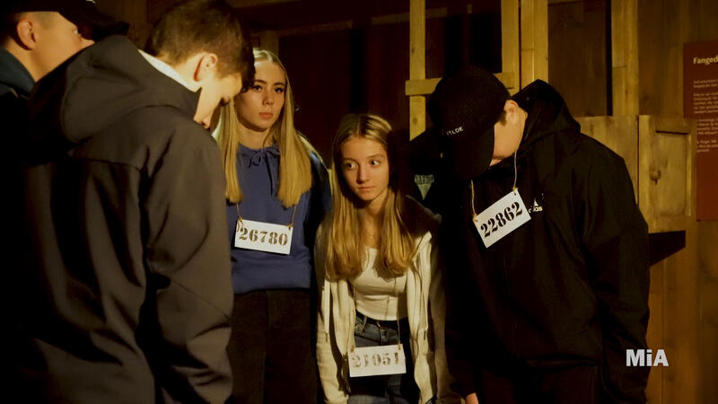 En gruppe ungdommer, med nummerskilt rundt halsen, står oppstilt foran køyesenger.