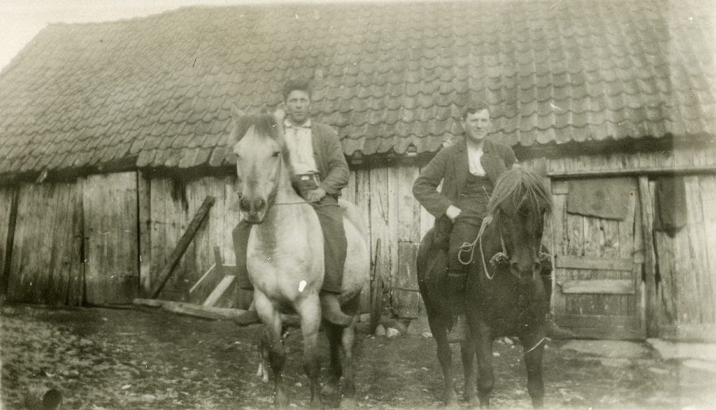 To menn sit på hest med ei løe i bakgrunnen.