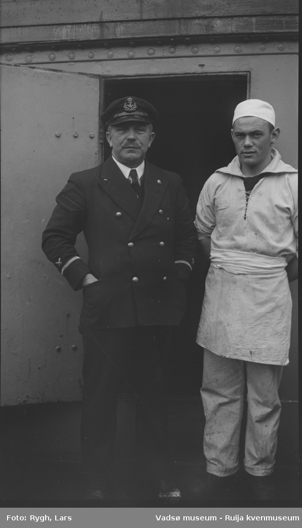 To sjømenn avbildet foran en skipsdør. De har ulike sjøuniformer på seg. Bildet har tilhørt Lars Rygh, og er tatt i forbindelse med minerydding i Varangerfjorden 1917 - 1918.
