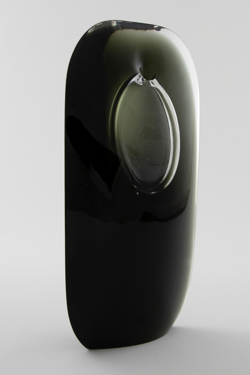 Skulpturell vase i klart og farget glass, hvor sistnevnte er opakt med et mørkt grå-grønnlig skjær. Rektangulær hovedform med mykt avrundede hjørner samt svakt konveks for- og bakside. På øvre del en elipseformet luftboble i klart glass med en liten åpning. Planslipt bunn.