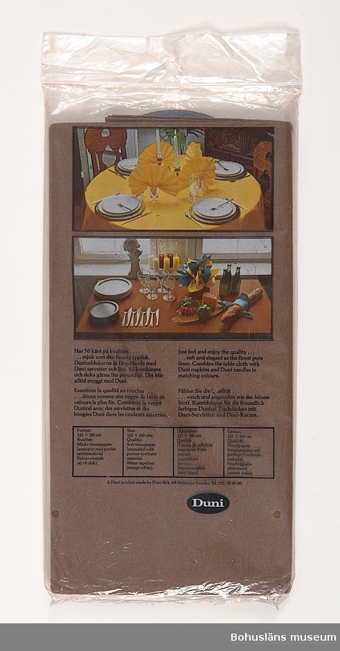 Bordsduk av brunt papper i originalförpackning, påse av plast med påtryck i färg och texten "DUNI Dunicel bordsduk 125 x 180 cm.
På baksidan foto på dukningsförslag av  middagsbord på gul resp. brun pappersduk med pappersservetter.