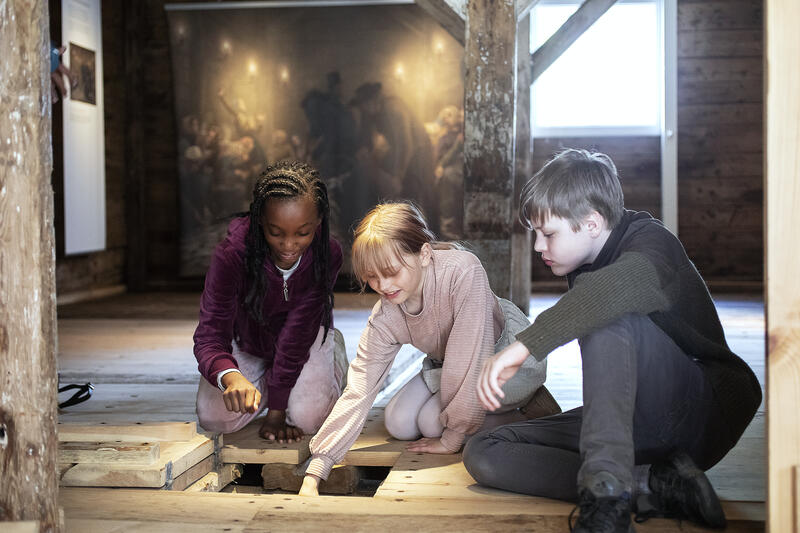 Tre barn sitter på gulvet og studerer luken i gulvet i silderommet på Norges Fiskerimuseum