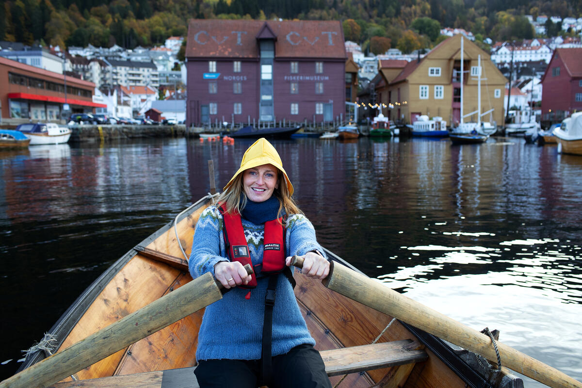 Vi ser en blid dame som sitter med redningsvest og gul sydvest og ror en tradisjonsrobåt mot Norges Fiskerimuseum