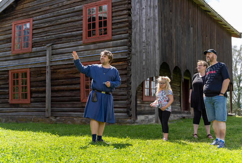 Guide i middelalderdrakt står foran et laftet, gammelt hus og peker og forteller til en familie.