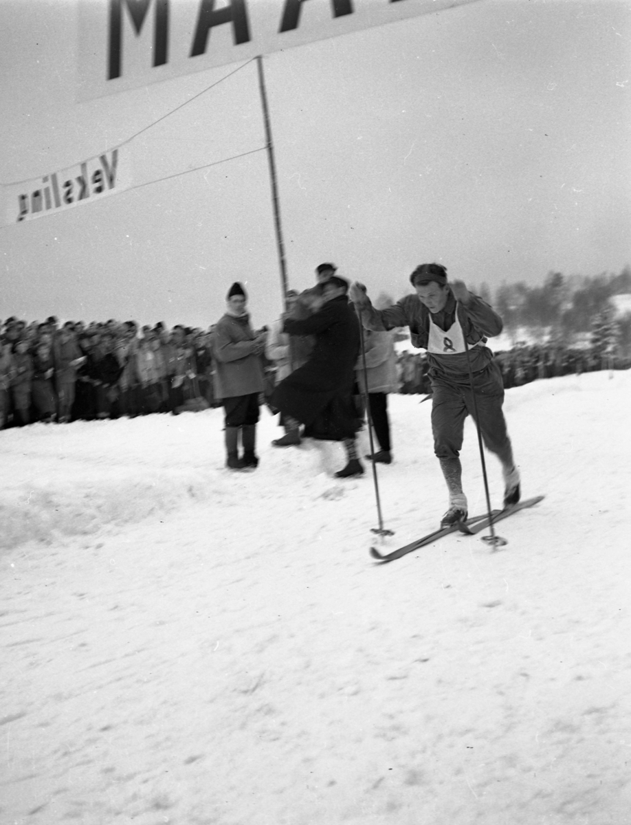 En skiløper med startnummer åtte i full fart i løypa. Mange tilskuere sees i bakgrunnen.