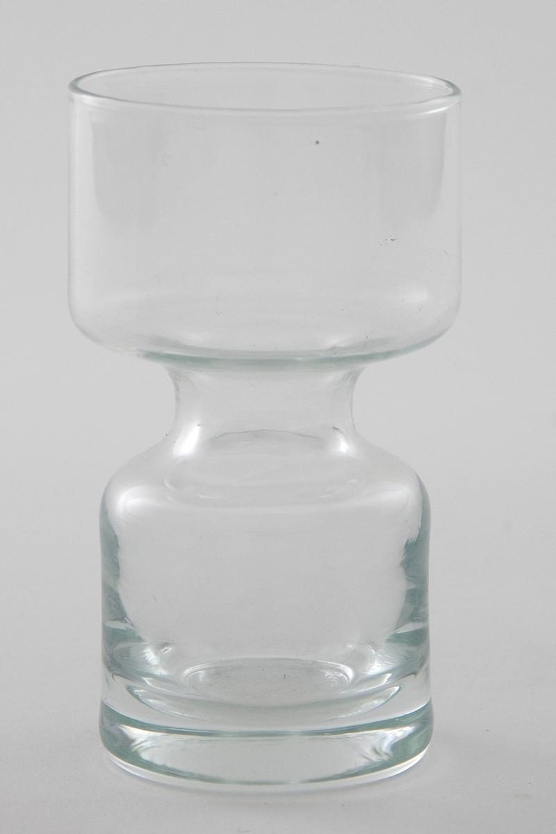 Lysestake i klart glass utformet som en sylinder som snevres inn på midten. Den øverste delen er litt videre enn den nederste.