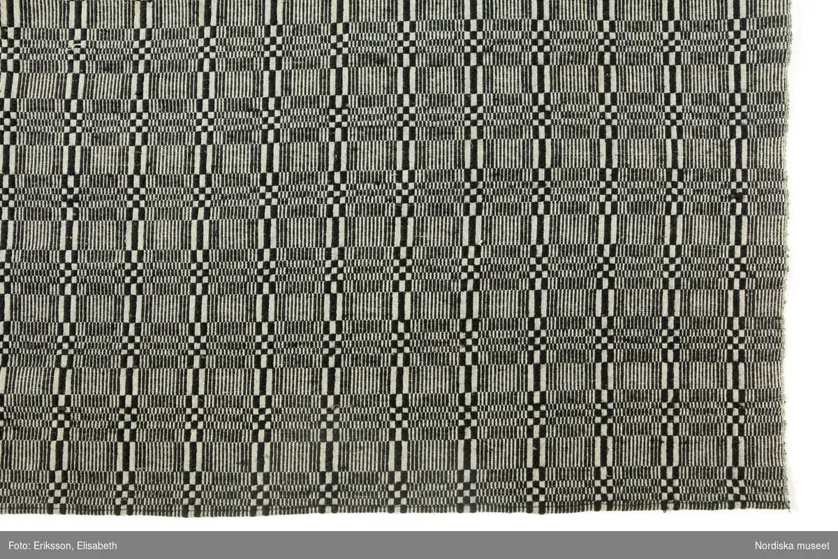 Täcke, varp av z-tvinnat vitt bomullsgarn, 44 trådar på 2 cm, inslag i botten av 2 trådar z-tvinnat grönt bomullsgarn och i mönstret 1-tr. s-spunnet svart ullgarn, förenklad dräll i rutmönster, vävt i 2 våder hopsytt på mitten för hand, maskinsydda fållar.

"Senast använt i en kyrkstuga i Nordmaling" ( 1922)
Linneband med mässingsringar ditsydda av museet för utställning.
Berit Eldvik april 2005