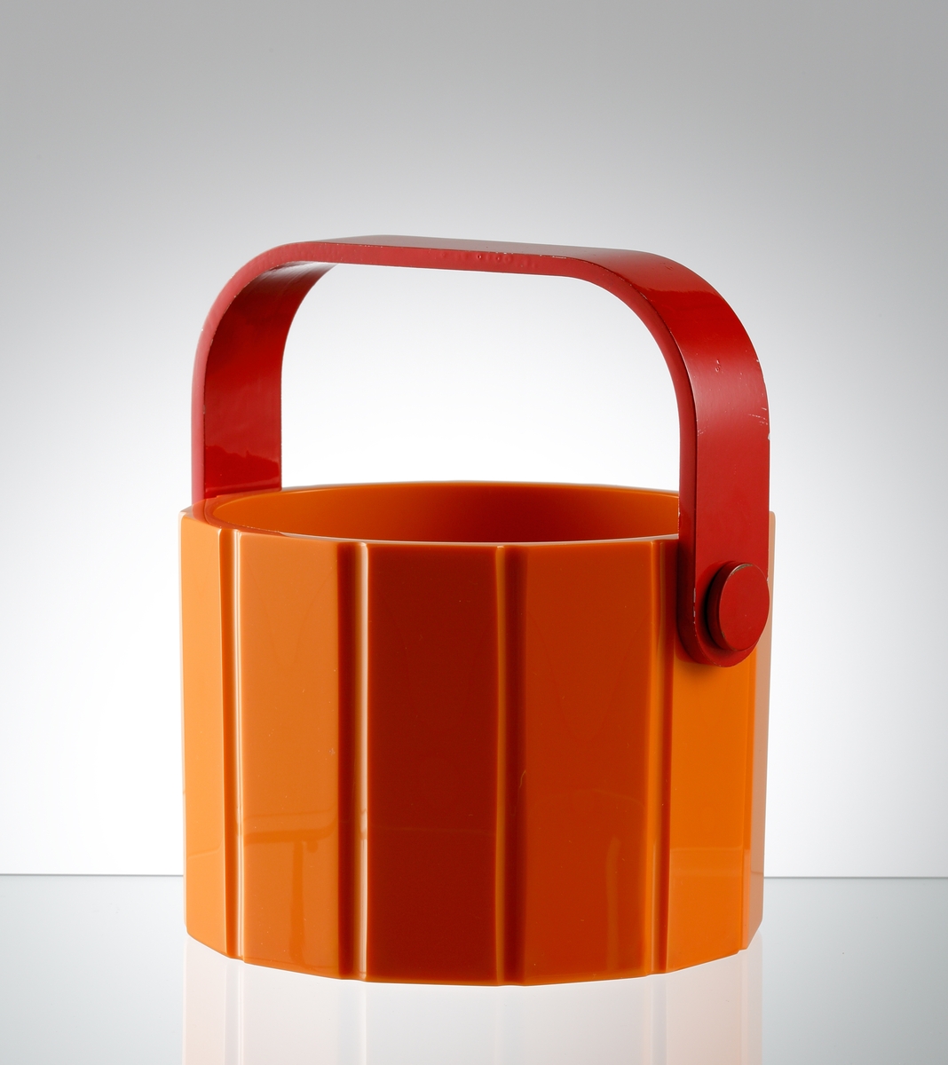 Formgiven av Jan Johansson. Orange glasmassa. Cylindrisk insida, tolvkantig utsida med stående räfflor. Rödmålad hänkel i metall, aluminium.