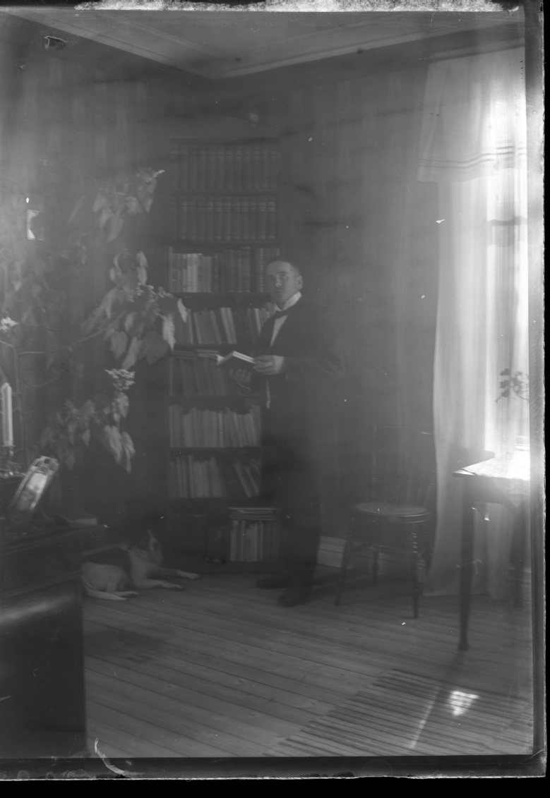 En man står med en bok i händerna vid en bokhylla full av böcker. Solen skiner in från höger.