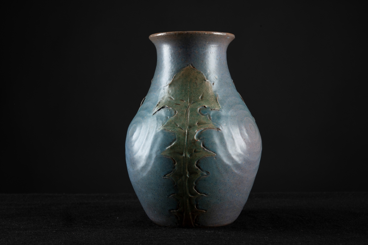 Jugenddekor i form av gröna maskrosblad som ligger tryckta mot vasens form från fotringen upp till två centimeter från mynningen, återkommer tre gånger runt om vasen. Blå bottenfärg med cirklar.