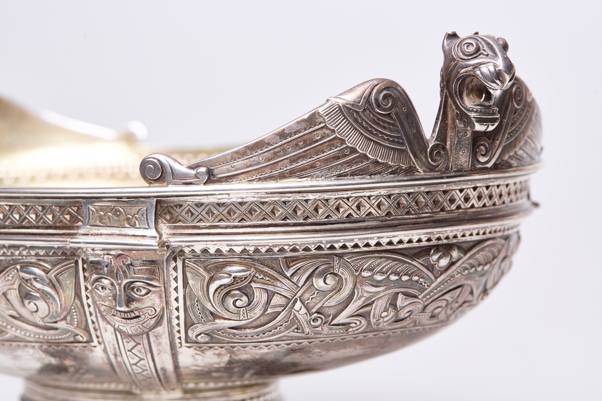 Stor, oval sølvbolle i form av et vikingskip med gravert norrøn dekor. Dragemønster, ansikter, dyrehoder. Fire føtter formet som dyrehoder. Dragestil.