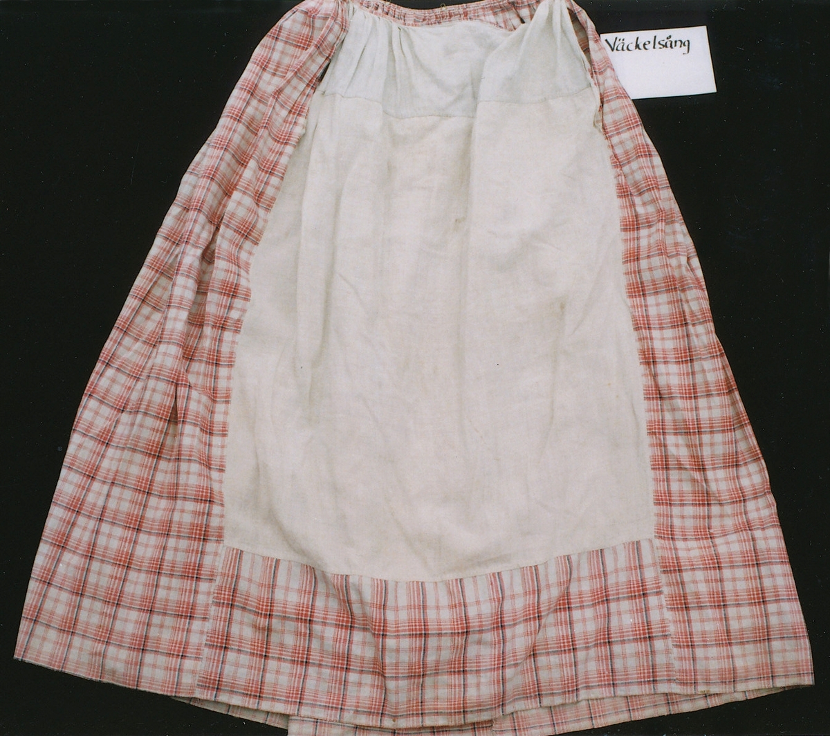 Tvådelad klänning med tröja och kjol i samma tyg storrutig bomullslärft i rött och blått på vit botten. 
Liv: Kort modell med uppvikt nederkant. Två framstycken/sidostycken dubbelknäppning med 3 par klädda knappar, tränsade knapphål på vänster sida, två små utåtlagda slag vid halsringningen. Ett bakstycke avsmalnande neråt med två skörtflikar, den högra iskarvad. Nederkanten infodrad med tyget och uppvikt tillsammans med de två skörtflikarna.
Lång ensömsärm, vid upptill med små veck på ärmkullen, avsmalnande mot handleden. 
Foder i bakstycke och höger framstycke av smårutig linnelärft i blått, brunt och gult på vit botten, i vänster framstycke och i ärmarna av blekt linnelärft. Bak i fodret 2 hakar att fästa mot kjolen.

Kjol i tre våder plus en iskarvad sparvåd av blekt linnelärft framtill som doldes under förklädet. Sparvåden är slät mitt fram, mot sidorna små lagda veck, 24 cm långa sprund/öppning vid var sida.  Våderna är tätt veckade mot smal midjelinning av tyget som knäpps i vänster sida med hyska och hake, baktill på linningen 2 hyskor att fästa mot tröjan. 
Smal fåll utan skoning i nedertkanten.

/Birgitta Blixt 2017