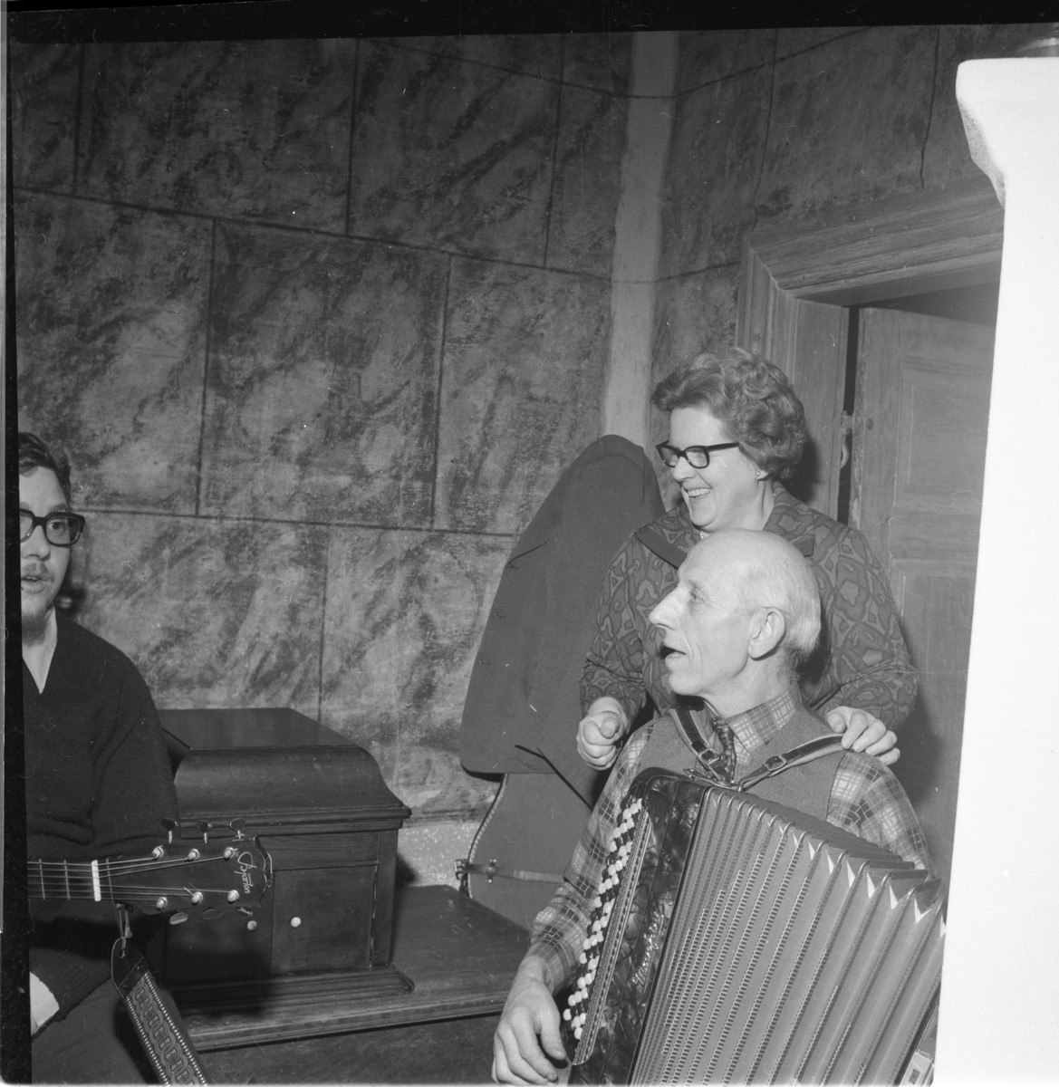 En man spelar dragspel, en kvinna står bakom honom och skrattar. En med man gitarr skymtar till vänster. Det är Ölmstad hembygdsförenings julfest.