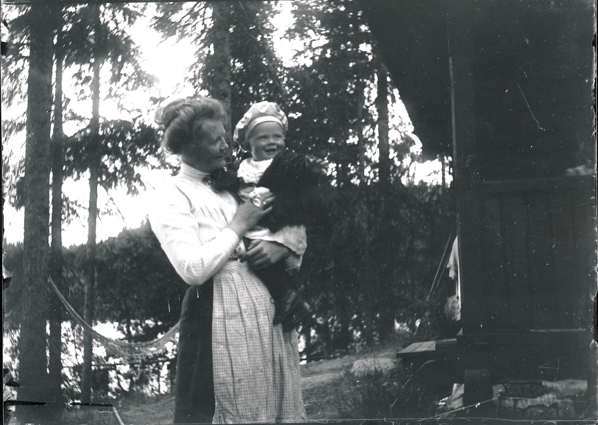 Nicoline Rydgren holder yngstesønnen, Wilhelm Fredrik, på armen. De står utenfor Mosjøhytta med Mosjøen i bakgrunnen.