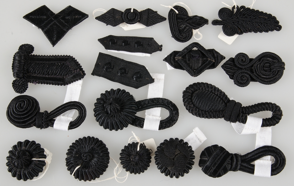 Diverse knappar, knapphål och s. k. flugor, trekantiga bitar av silke och siden. Halvfabrikat till knapptillverkning. De flesta svarta några kulörta. Samt diverse snörmakerier i svart silke och två band i snörmakeriarbete av svart silke.