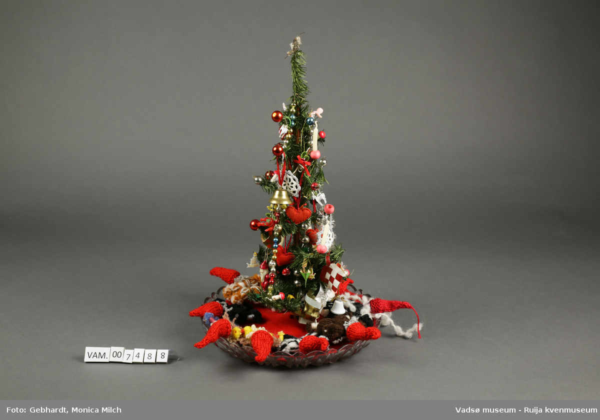 Lite juletre i plast. Juletreet er pyntet med stearinlys, bjeller, julekuler, nisser, julekurver og annen tradisjonell julepynt. Treet står på et lite glassfat med ei rød filtduk på. Rundt treet er det plassert ni garnnisser i ulike farger.