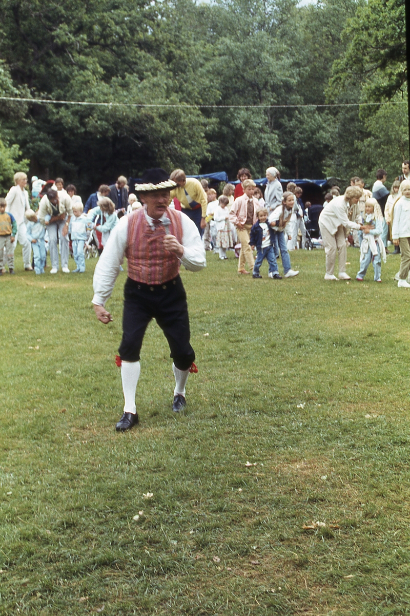 Barn och vuxna dansar kring stången vid midsommarfirande på Ekensås cirka 1982. Lekledare är Sven Olof "Olle" Svensson (klädd i folkdräkt).