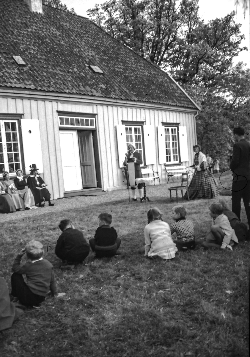 Utkledning og rollspill ved hovedhuset på Berg gård,1970-tallet.
Bjørn Sandberg sittende ved hovedinngang og Inger Sandberg, stående til hoyre. Folk i parken.