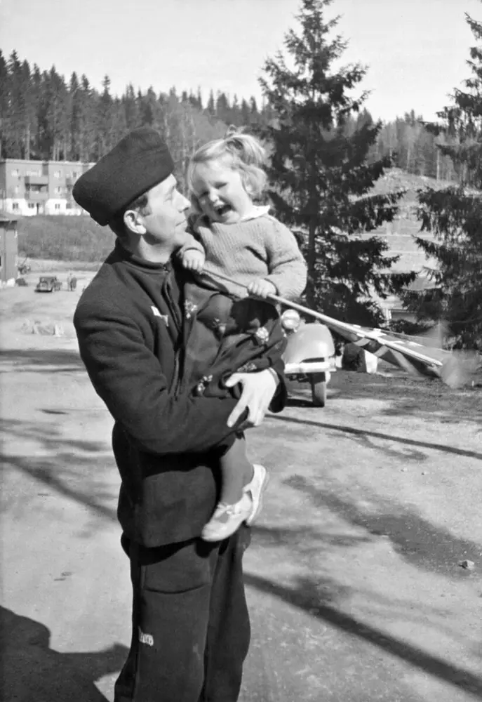 Photo from Grini prison camp 8. may 1945.  Black and white. Man in prison uniform carrying his toddler daughter. The daughter is holding a norwegian flag