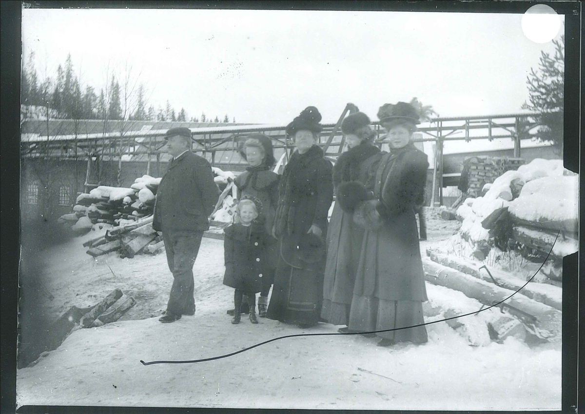 Medlemmer av familien Rydgren og fru Berger på tur på fabrikkområde. Fra venstre bestyrer Wilhelm Rydgren, Inger Elisabeth som står foran Wilfried, fru Berger, fru Nicoline Rydgren og Signe. I bakgrunnen er det synlig en kjerrat eller transportør og en fabrikkbygning.