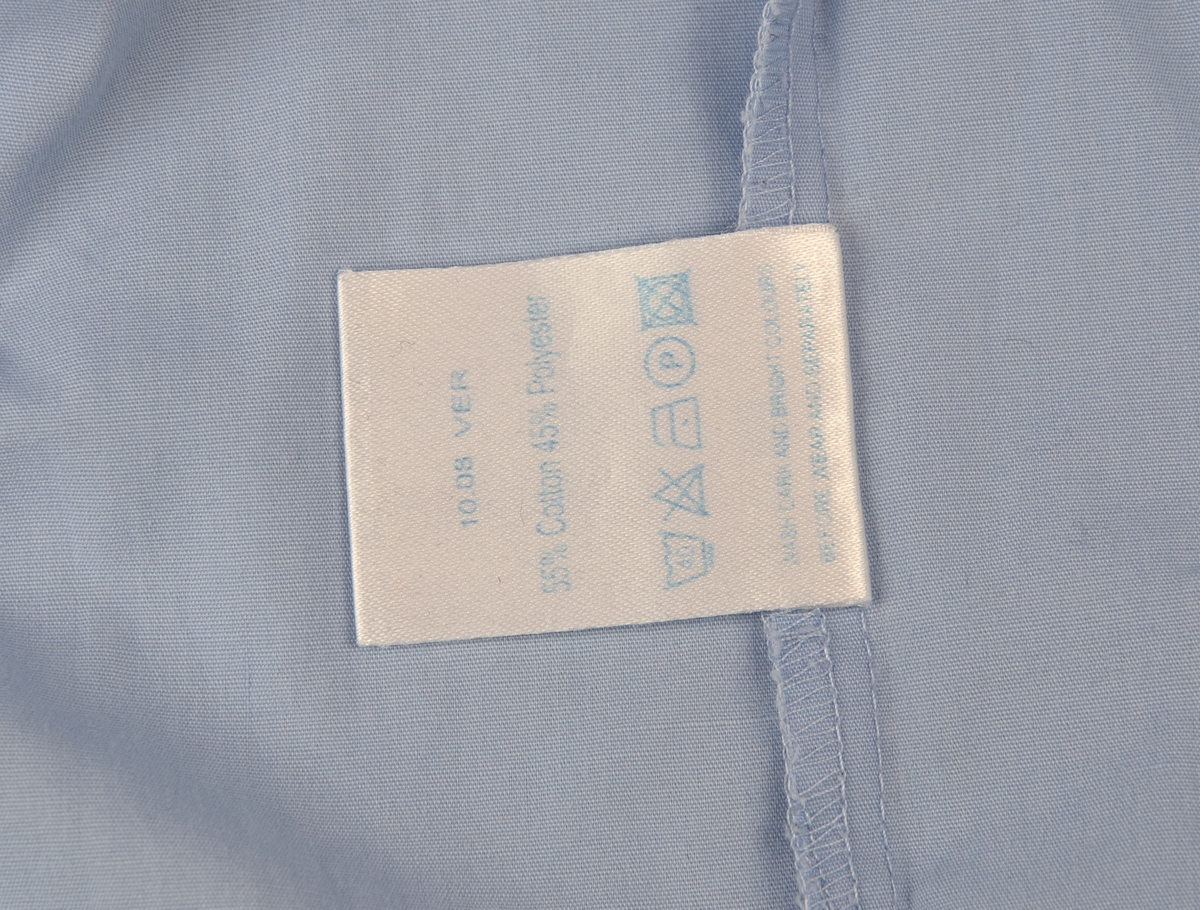 Ljusblå kortärmad skjorta i storlek 50. Skjortan är gjord i 55% bomull och 45% polyester. Knäpps med sju ljusblå knappar av plast på framsidan, två extra knappar finns längst ner på insidan av skjortan. En bröstficka på vänster sida. På vänster skjortärm finns TUI:s logotyp broderad i rött. En etikett i nacken där det står "World of TUI designed by GMK EUR 50". En tyglapp på insidan av skjortan med information om material samt tvättråd.