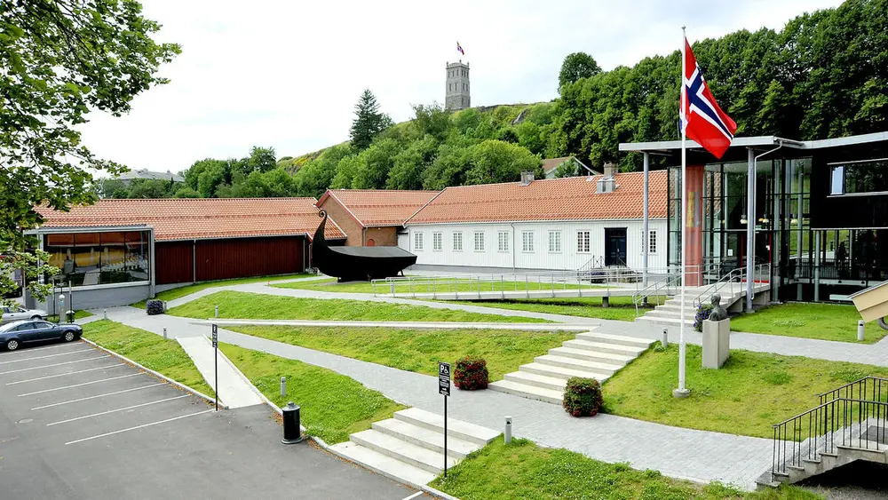 Bildet viser noen bygninger som ligger nedenfor en grønn ås der det står et murtårn helt øverst. Foran i bildet ser vi  litt plen og en trapp opp til inngangen. Det er en flaggstang med det norske flagget utenfor inngangen.