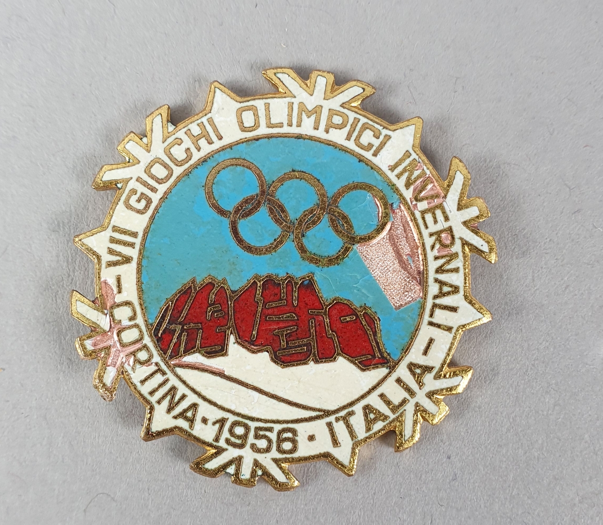 Rundt merke med hvit kant med gullbokstaver. De olympiske ringer i midten på blå bakgrunn over røde fjell og hvit snø