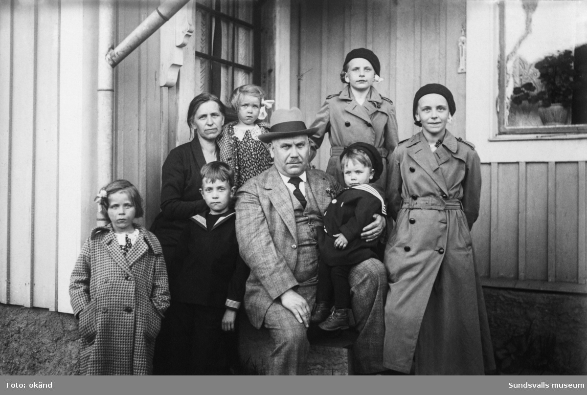 Familjeporträtt med ett par och sex barn framför ingången till ett bostadshus i trä. Ur en samling som tillhört Oscar och Greta Carlsson i Selånger.