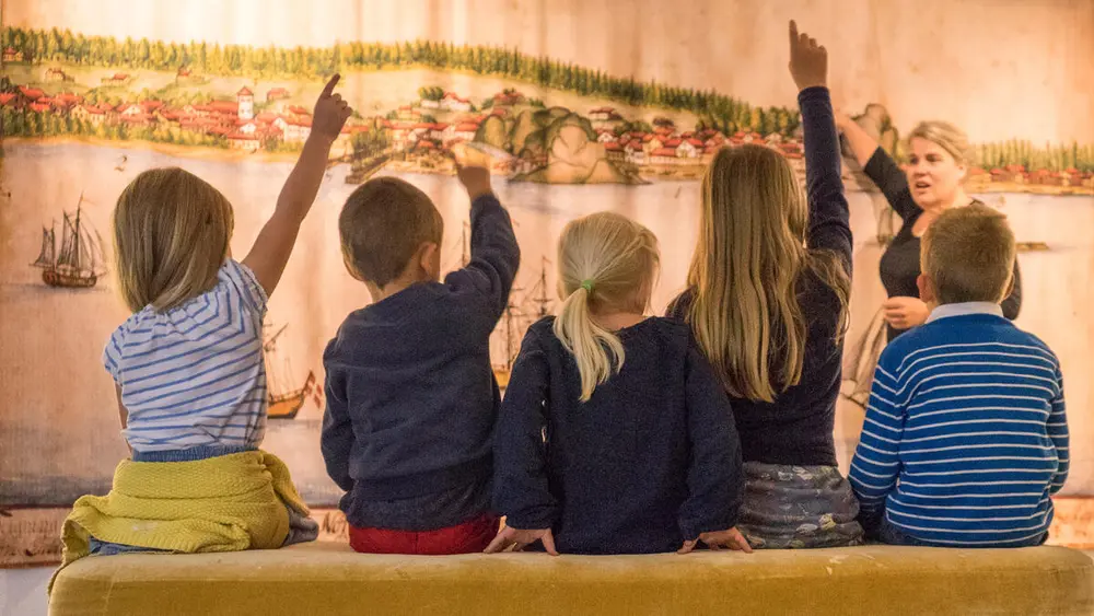 Bildet viser ryggene til barn som sitter på en benk. Mange av dem har en hånd i været. De ser på et stort maleri og en kvinne som står og snakker.