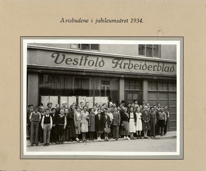 Et sort-hvitt foto er limt på en papplate. Bildet viser en gruppe mennesker som står oppstilt foran et bygg med skriften Vestfold Arbeiderblad på veggen. (Foto/Photo)